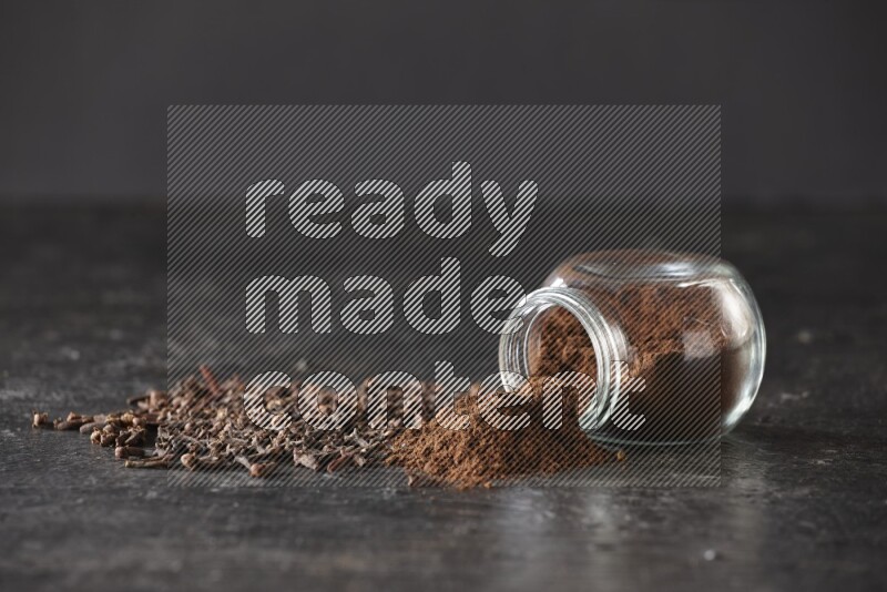 وعاء زجاجي للبهارات مقلوب ممتلئ ببودرة القرنفل والبودرة سقطت منه مع حبوب القرنفل المتناثرة على أرضية سوداء