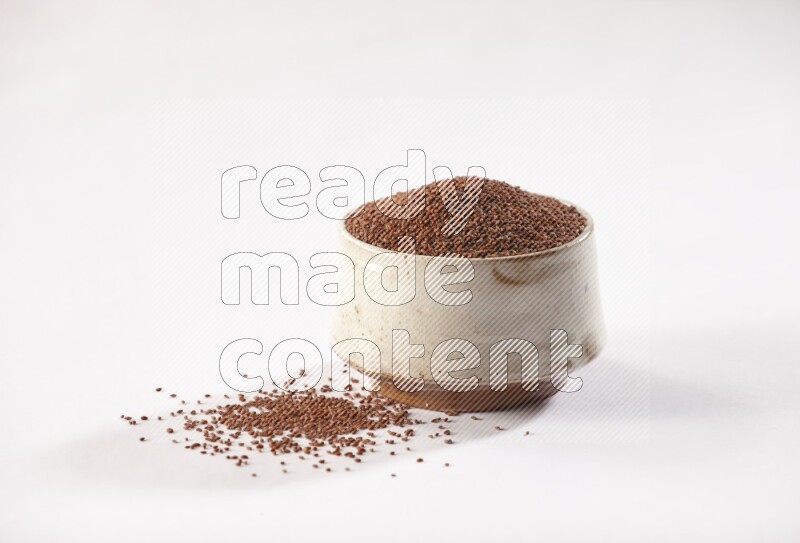 وعاء فخاري بيج ممتلئ ببذور حب الرشاد مع بعض البذور متناثرة على خلفية بيضاء