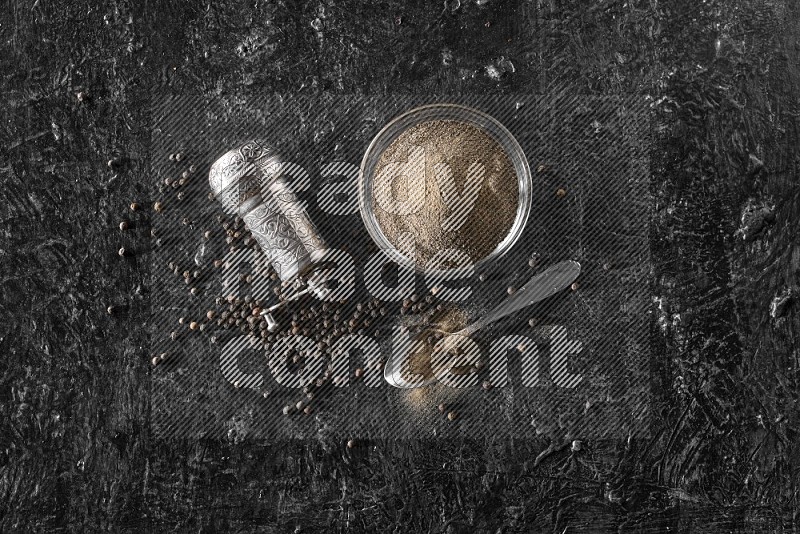 A glass bowl full of black pepper powder with black pepper beads, a turkish metal pepper grinder and a metal spoon on textured black flooring