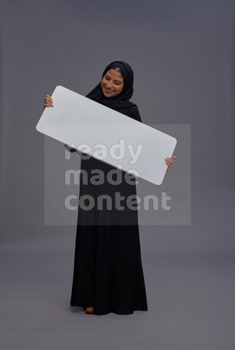 Saudi woman wearing Abaya standing holding white board on gray background