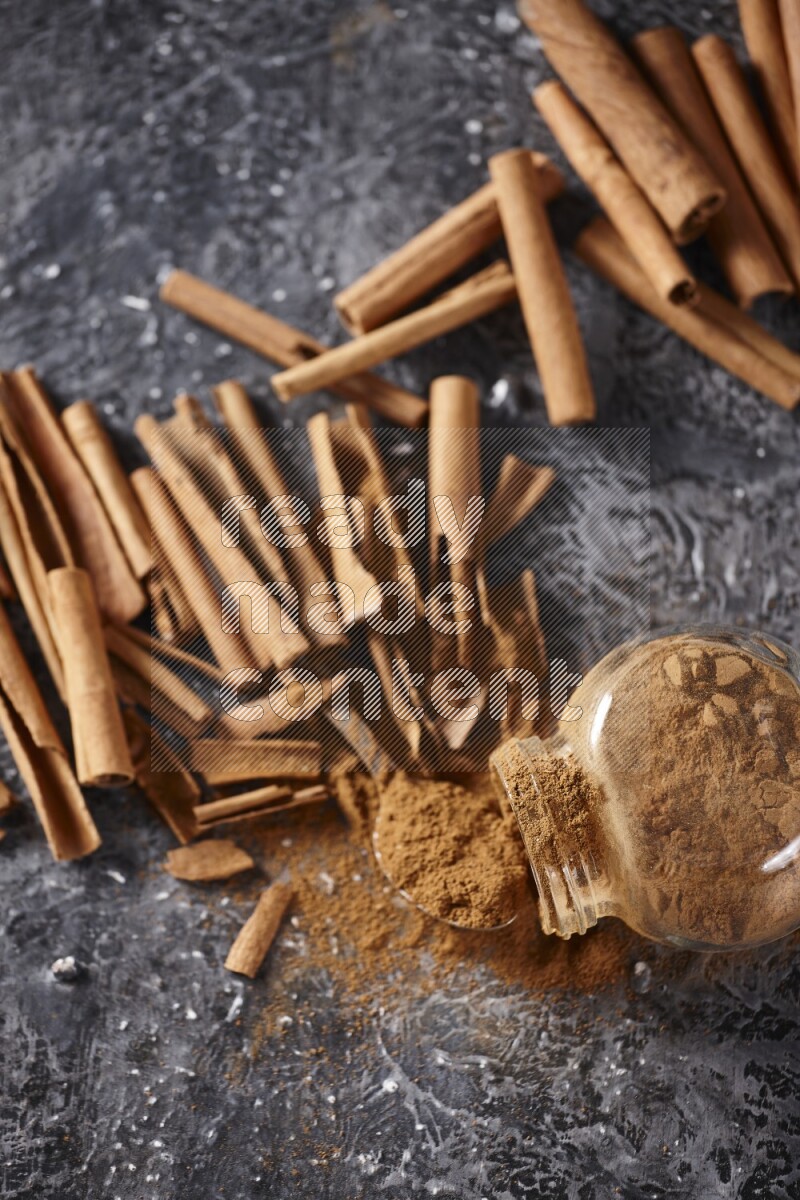 Herbal glass jar full cinnamon powder flipped and a metal spoon full of powder surrounded by cinnamon sticks on textured black background in different angles