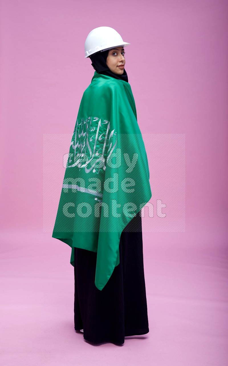 Saudi woman wearing Abaya with engineer vest and helmet standing holding Saudi flag on pink background