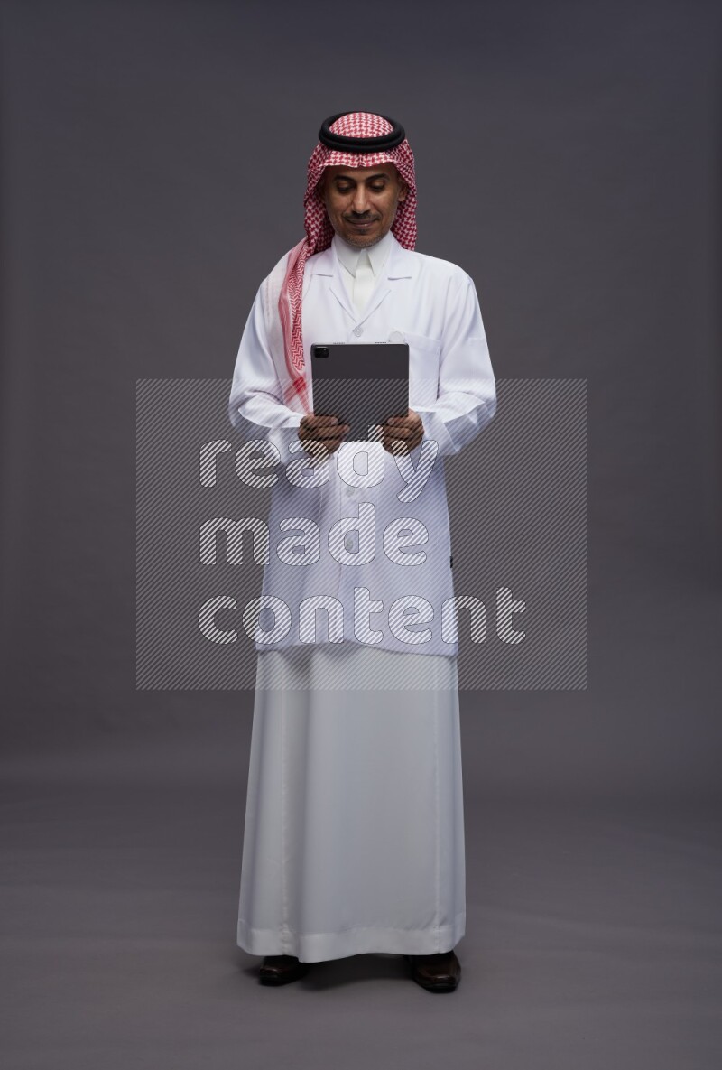 Saudi man wearing thob with lab coat and shomag with pocket employee badge standing working on tablet on gray background