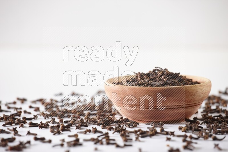 A wooden bowl full of cloves with spread grains on a white flooring