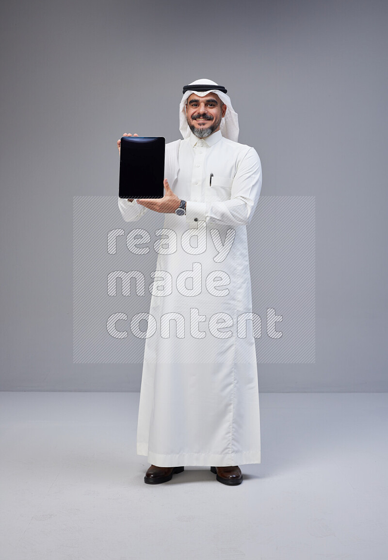 Saudi man Wearing Thob and white Shomag standing showing tablet to camera on Gray background