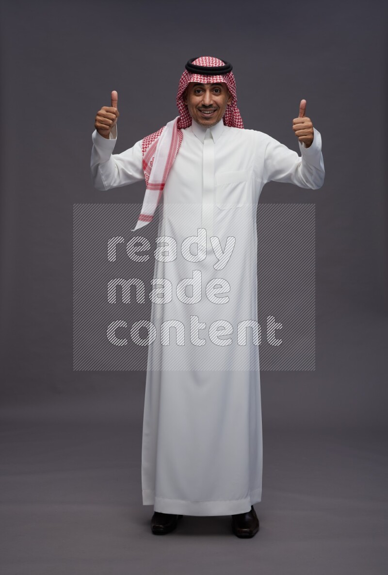Saudi man wearing thob and shomag standing interacting with the camera on gray background