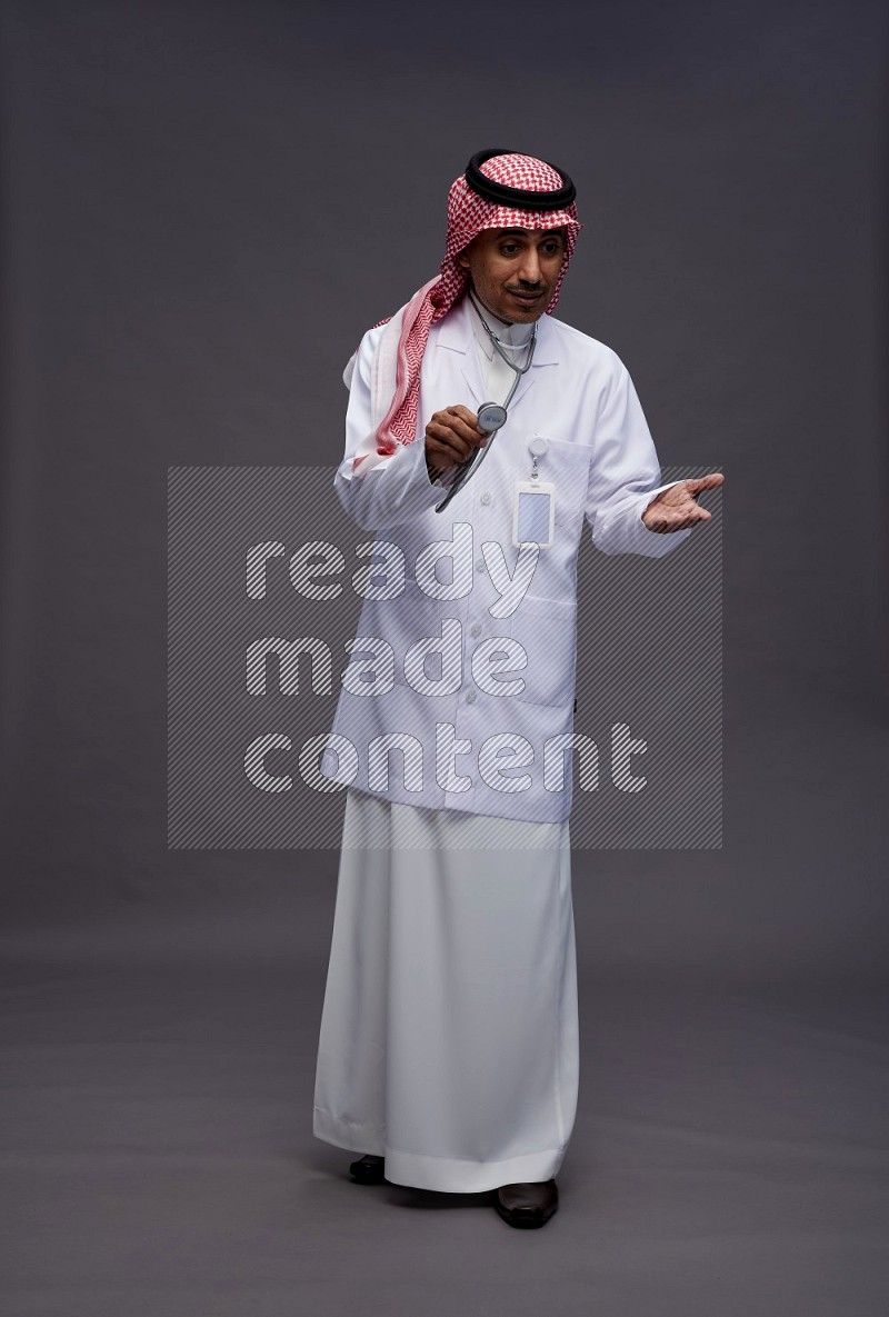 Saudi man wearing thob with lab coat and shomag with pocket employee badge with stethoscope standing interacting with the camera on gray background