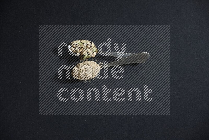 2 Metal spoons full of cardamom powder and cardamom seeds on black flooring