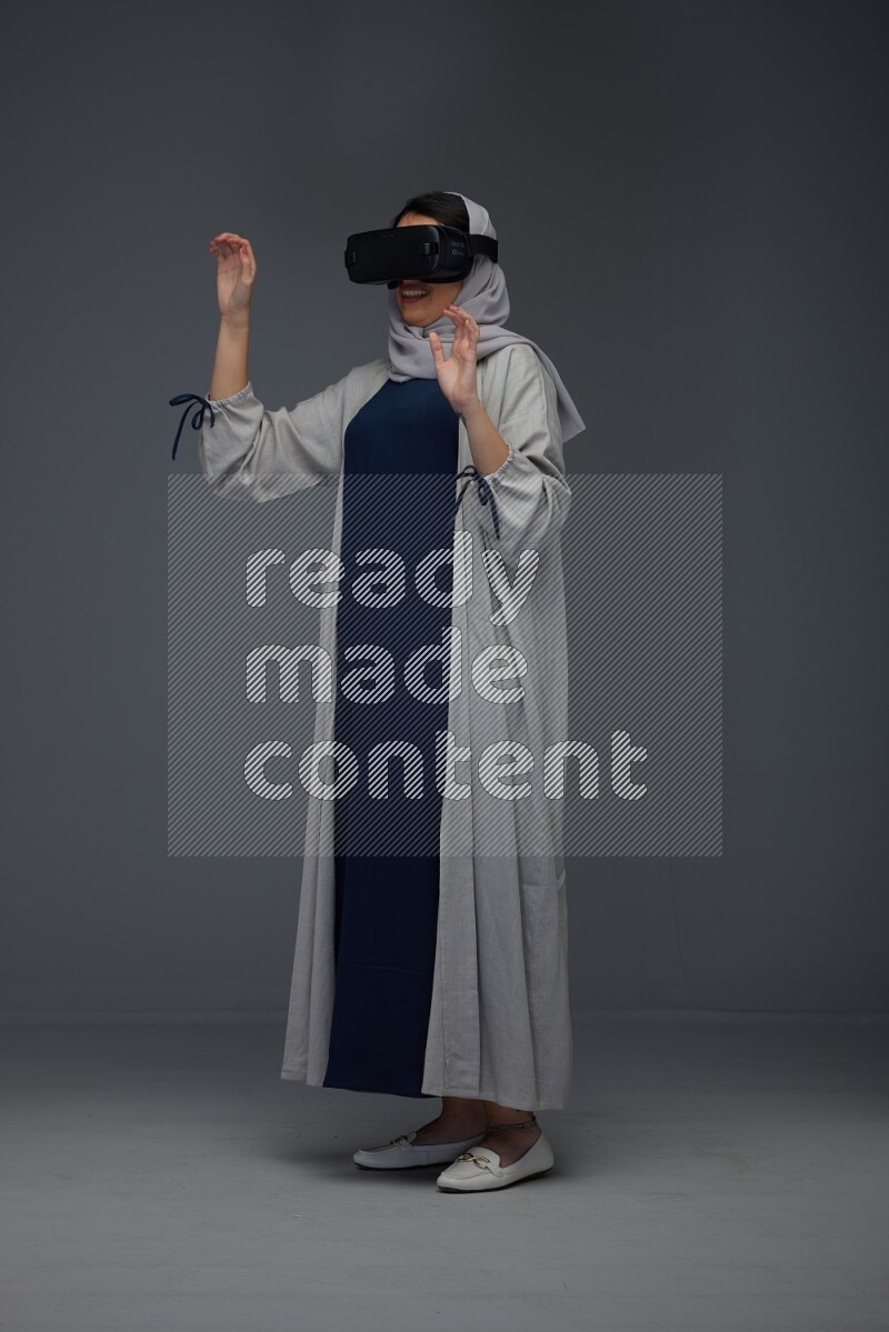 A Saudi woman wearing a light gray Abaya and head scarf standing and wearing VR eye level on a grey background