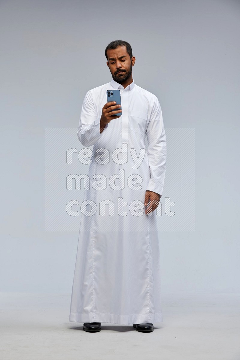 Saudi man wearing Thob standing texting on phone on Gray background