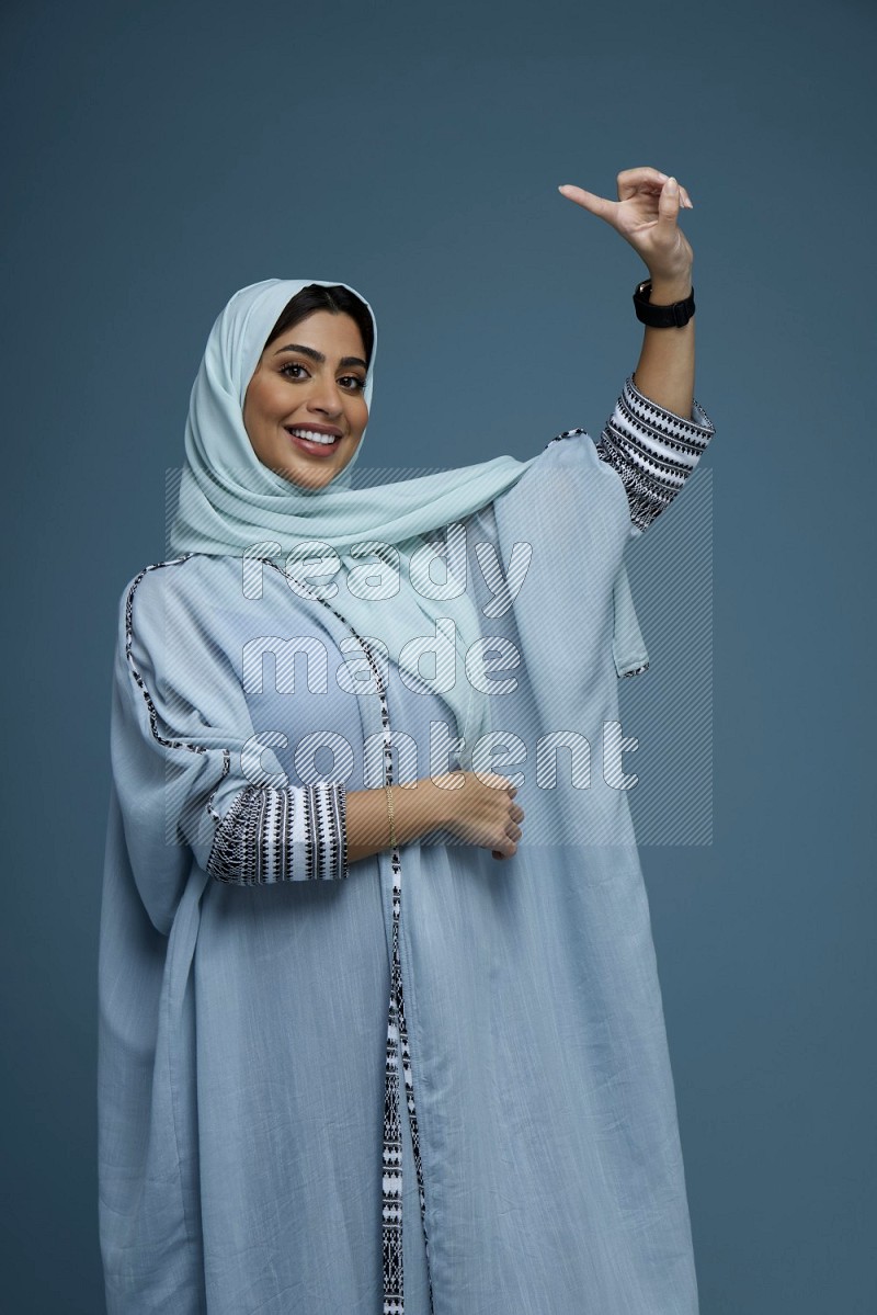 A Saudi woman pointing in a blue background wearing a blue Abaya with hijab