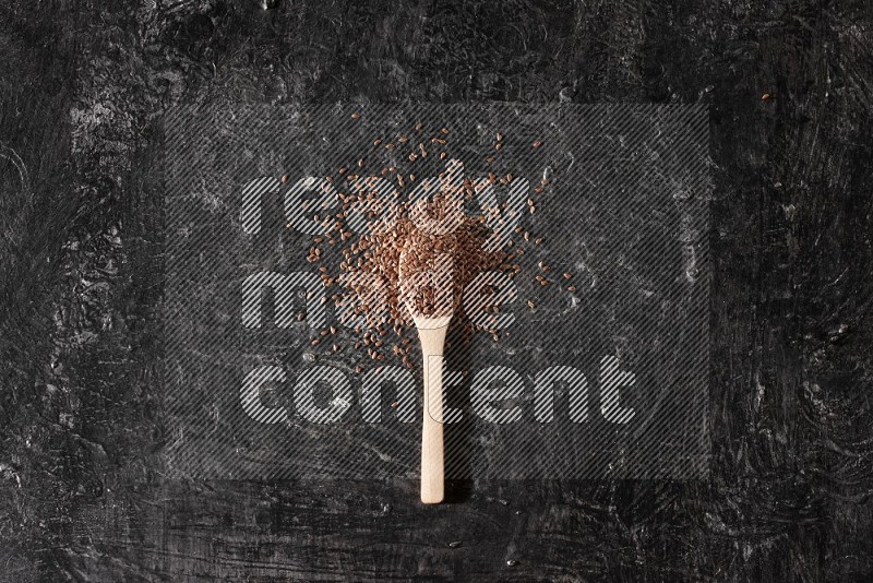 A wooden spoon full of flaxseeds and surrounded by seeds on a textured black flooring