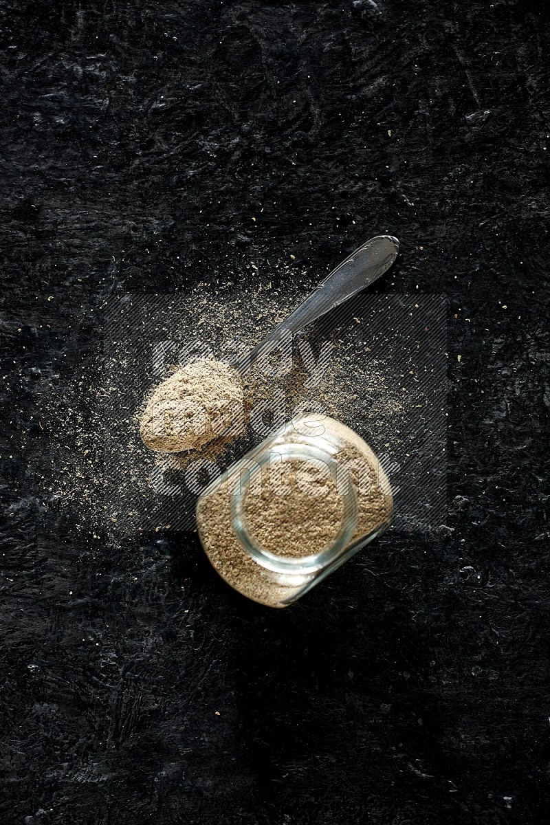 A glass spice jar and metal spoon full of cardamom powder on textured black flooring