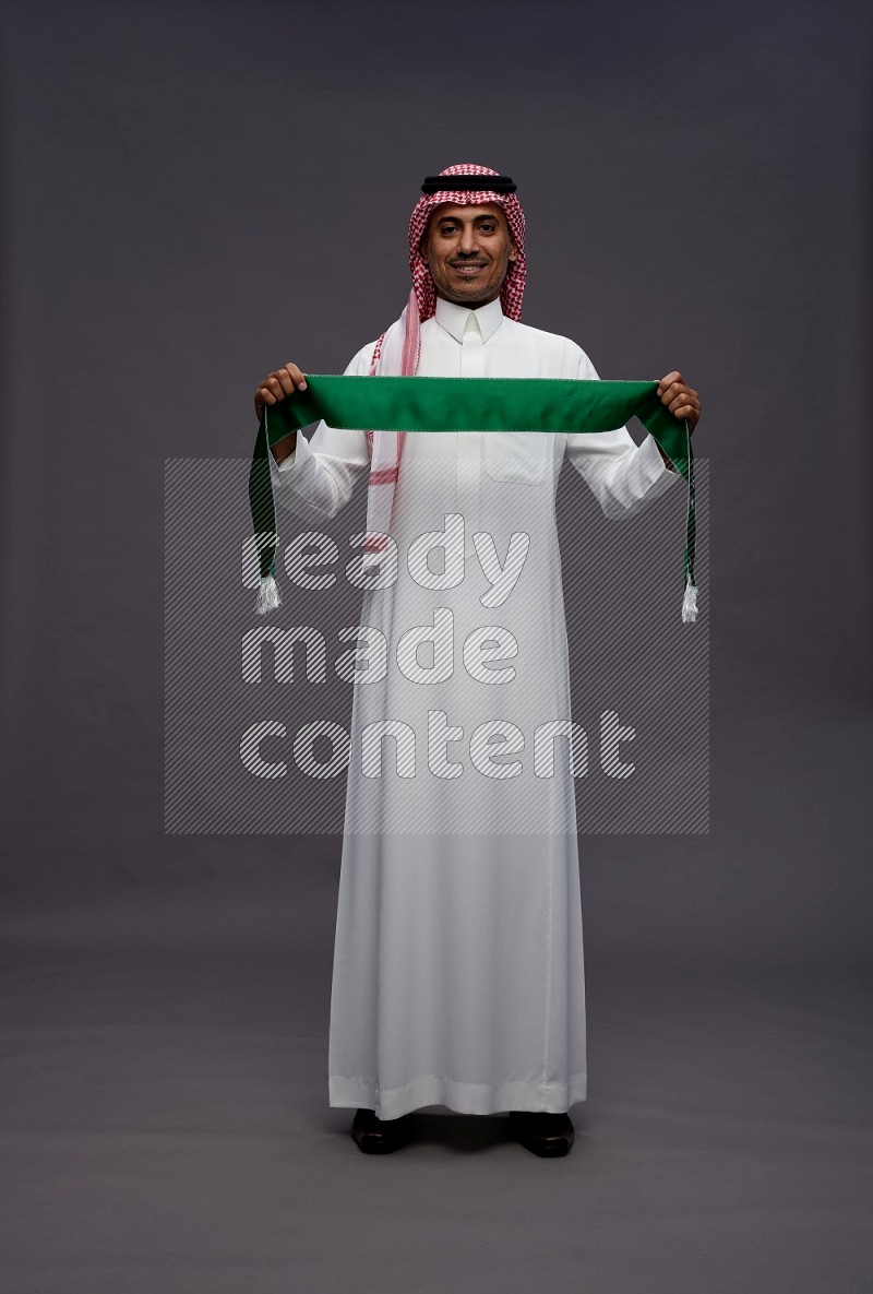 Saudi man wearing thob and shomag standing holding Saudi flag on gray background