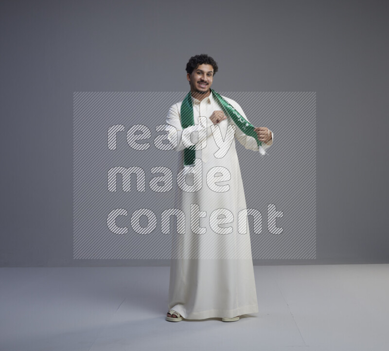 A Saudi man standing wearing thob and Saudi flag scarf on gray background