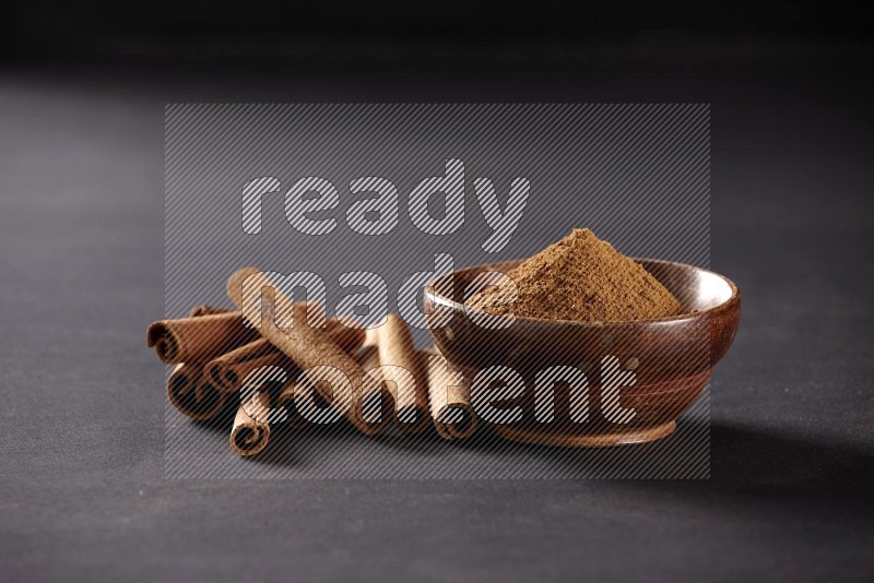 Cinnamon sticks stacked beside a wooden bowl full of cinnamon powder on black background