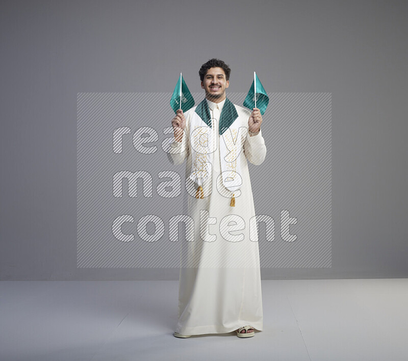 A Saudi man standing wearing thob and saudi flag scarf and holding small saudi flag on gray background