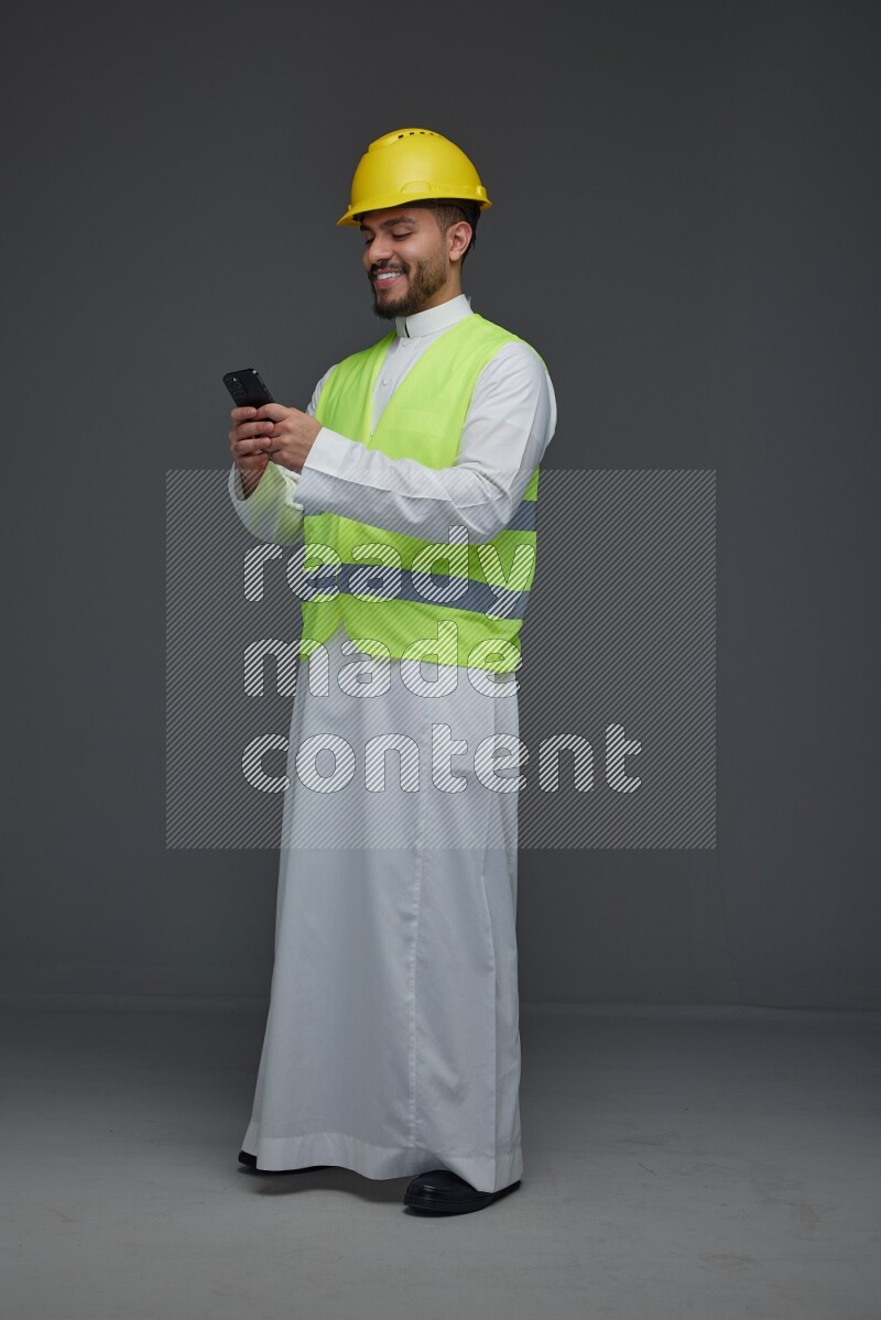 A Saudi man wearing Thobe with a yellow safety vest and white helmet standing and using his phone eye level on a gray background