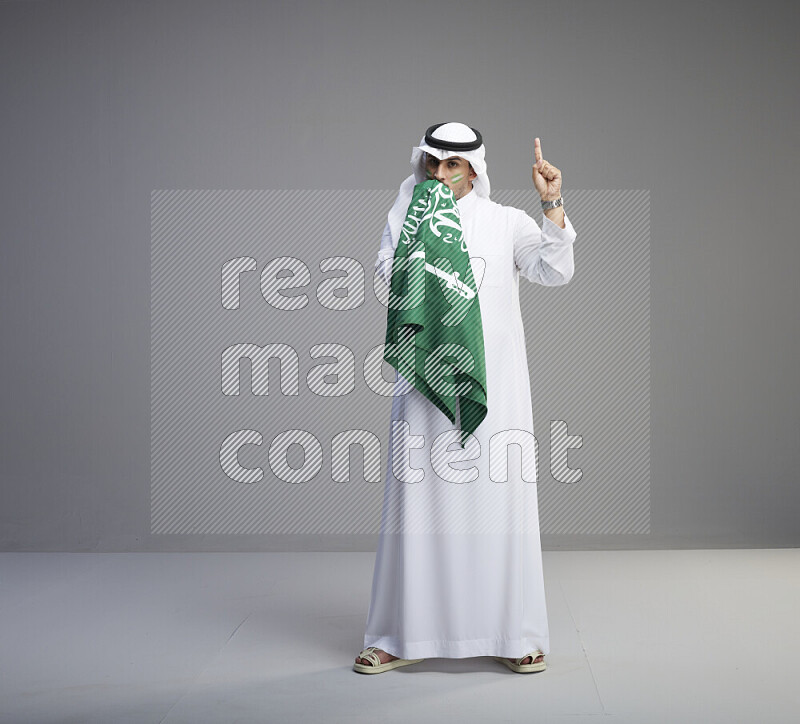 A Saudi man standing wearing thob and white shomag with face painting kissing big Saudi flag on gray background