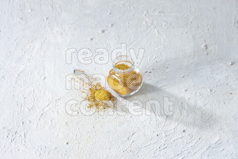 A glass spice jar and metal spoon full of turmeric powder on textured white flooring