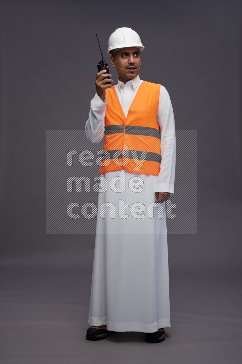 Saudi man wearing thob with engineer vest standing holding walkie-talkie on gray background