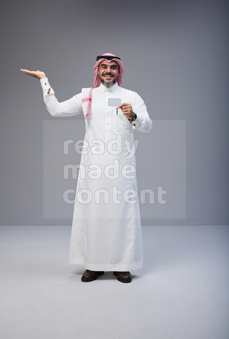 Saudi man Wearing Thob and red Shomag standing holding ATM card on Gray background