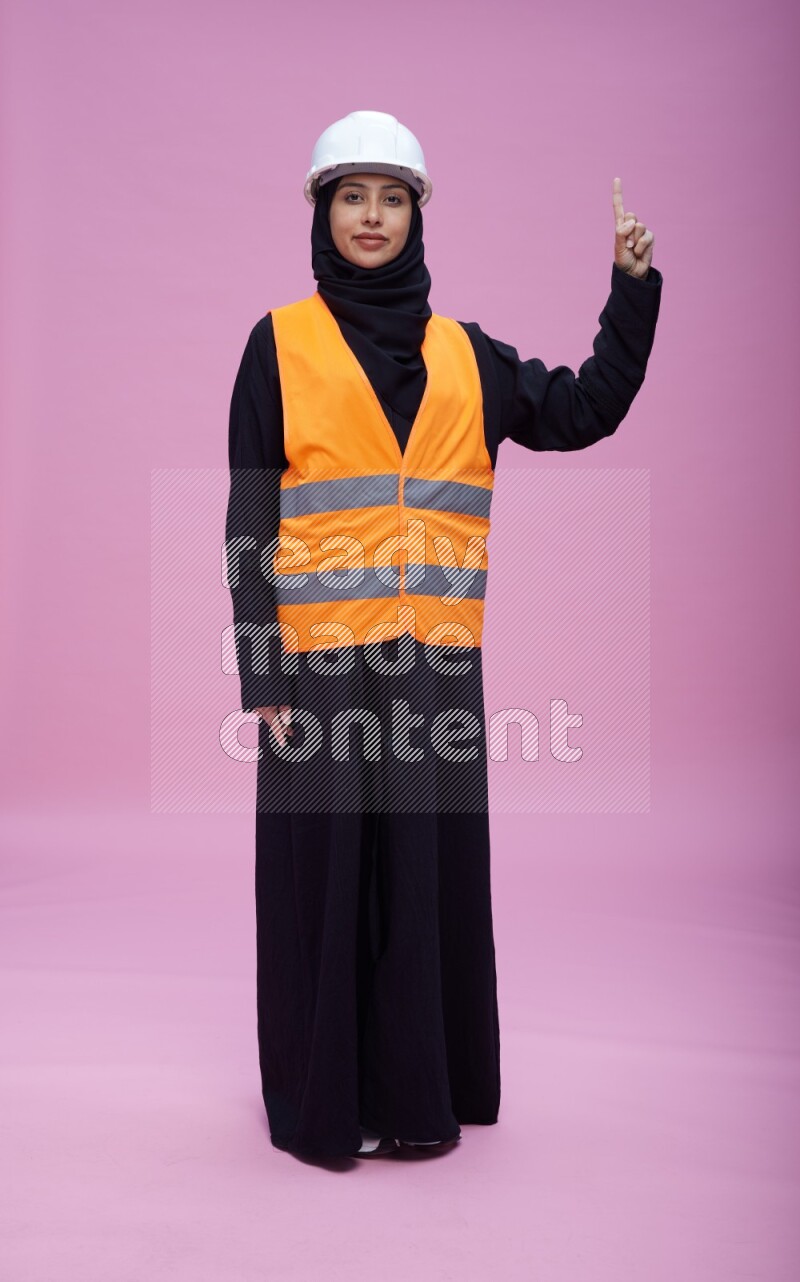 Saudi woman wearing Abaya with engineer vest and helmet standing interacting with the camera on pink background