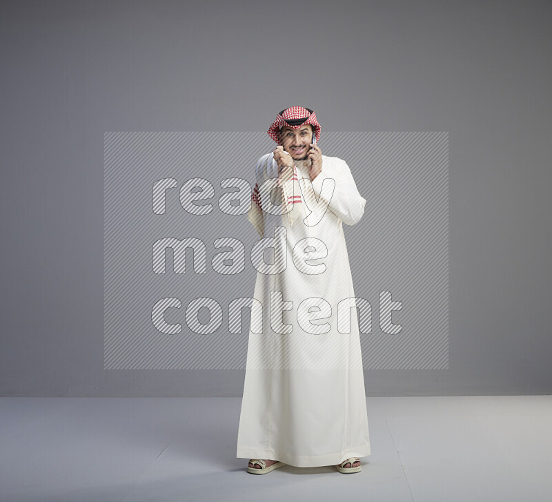 A Saudi man standing wearing thob and red shomag talking on phone on gray background