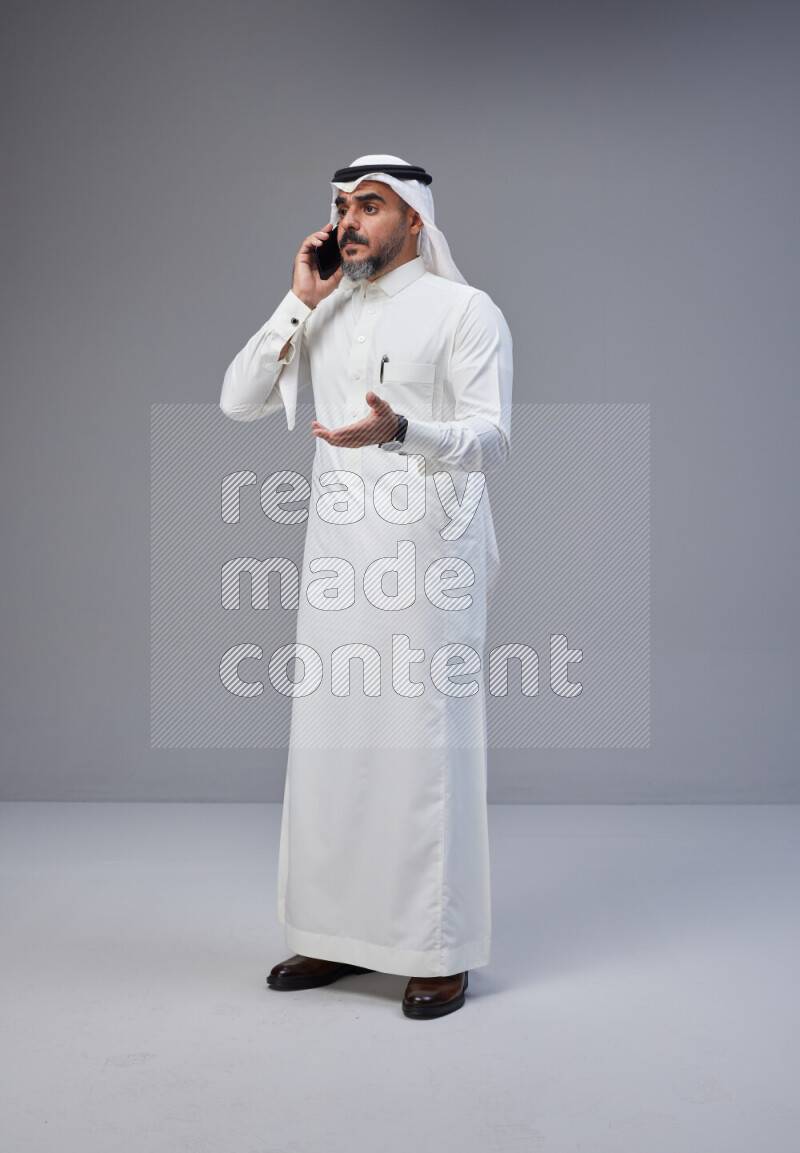 Saudi man Wearing Thob and white Shomag standing talking on phone on Gray background