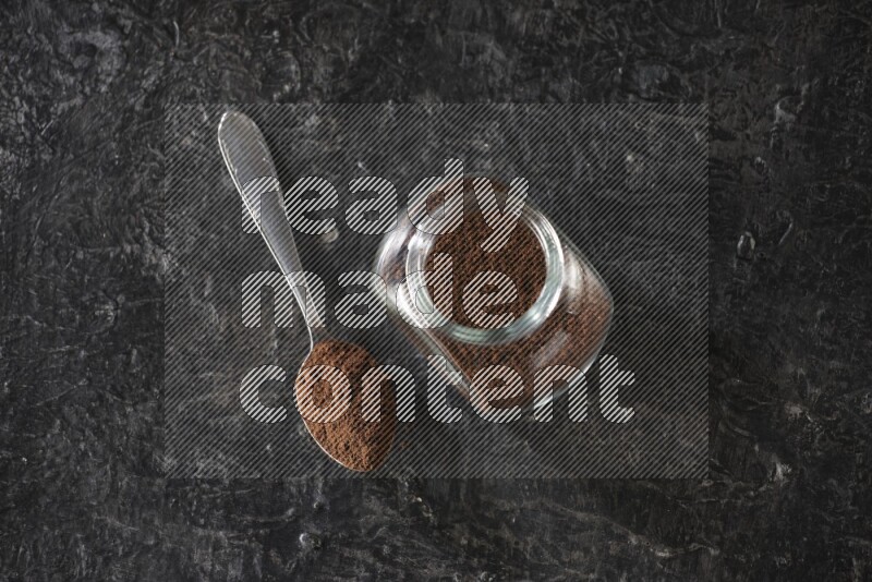 A glass spice jar and a metal spoon full of cloves powder on textured black flooring