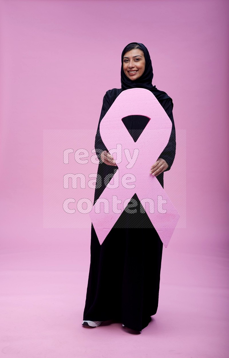 Saudi woman wearing pink ribbon on abaya standing holding awareness ribbon on plain pink background