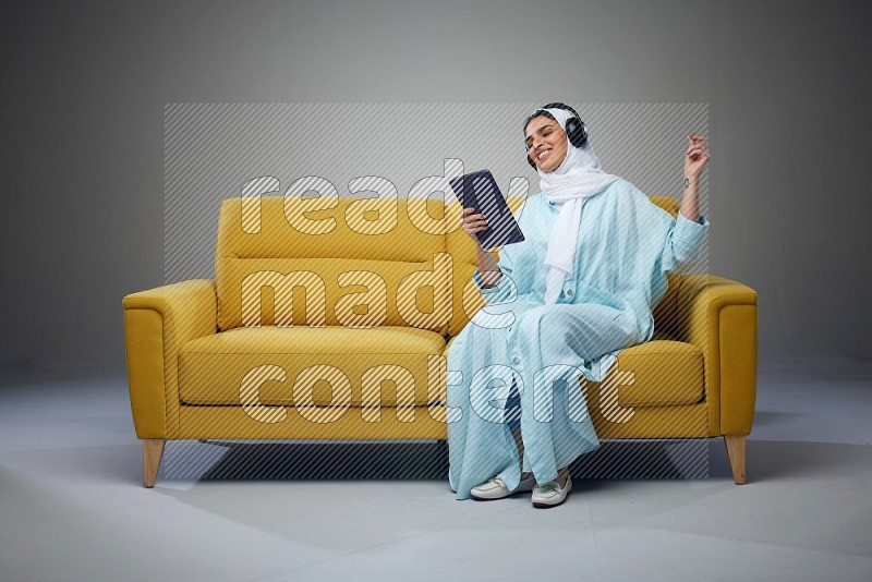 A Saudi woman wearing a light blue Abaya and a white head scarf sitting on a yellow sofa and holding an iPad while wearing headphone eye level on a grey background