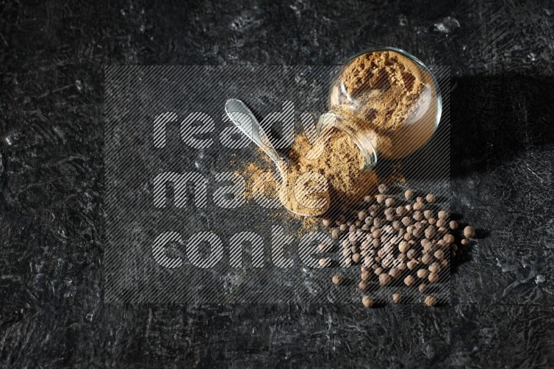 وعاء زجاجي للتوابل مقلوب وملعقة معدنية ممتلئان ببودرة بهار الفلفل الحلو والبودرة تناثرت من الوعاء مع بعض الحبوب على خلفية سوداء