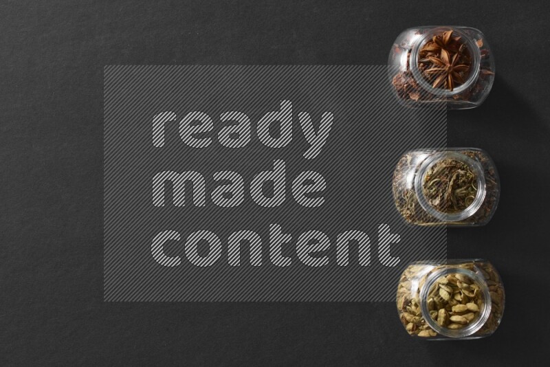 3 glass spice jars full of star anise, basil and cardamom on black flooring