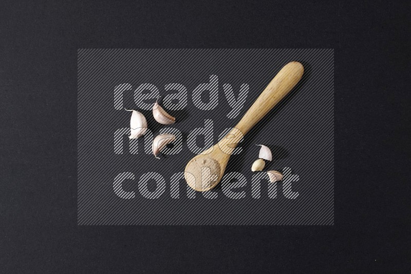 A wooden spoon full of garlic powder with cloves beside it on a black flooring in different angles