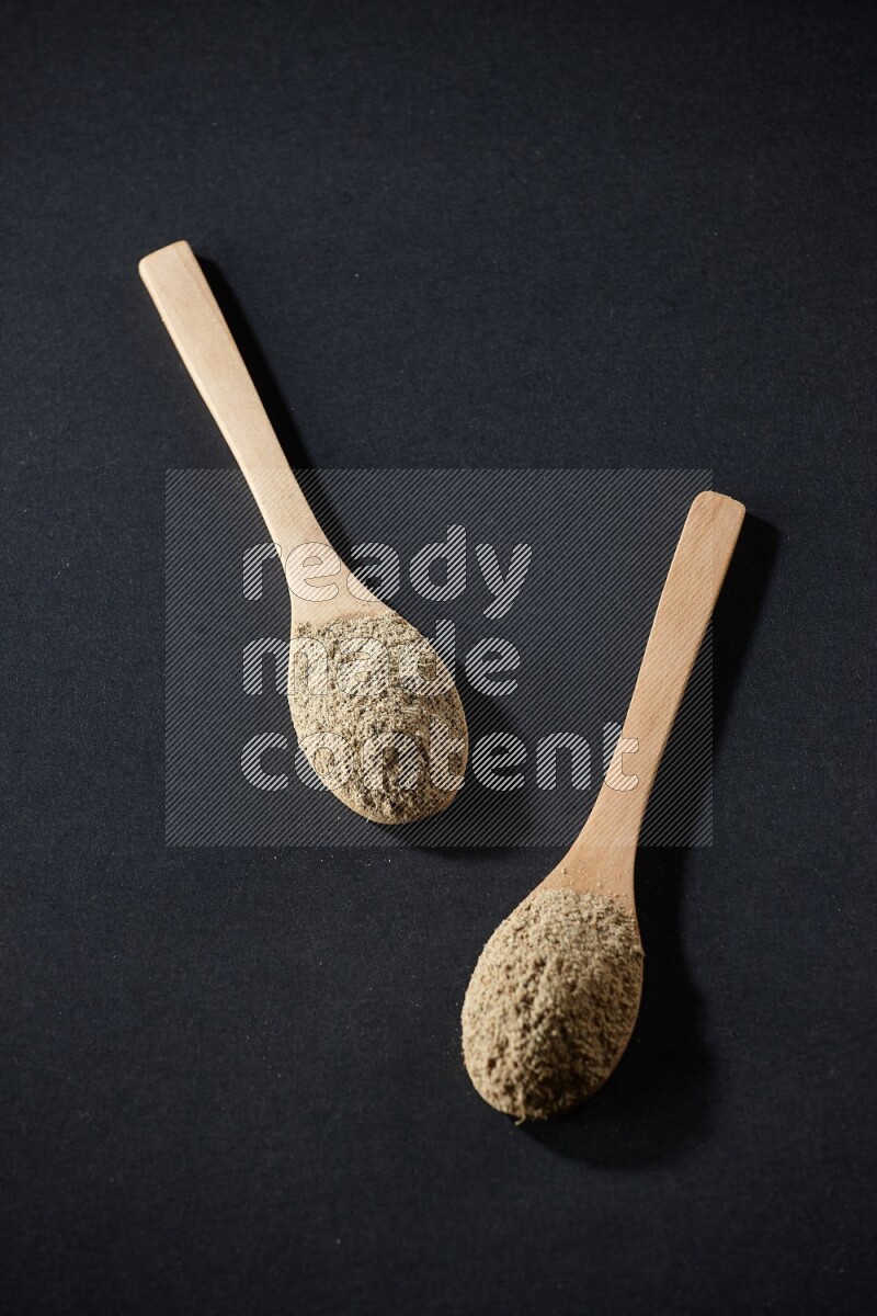 2 Wooden spoons full of cardamom powder on black flooring