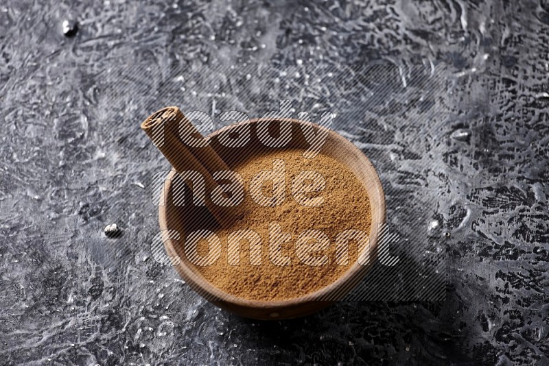 Wooden bowl full of cinnamon powder and a cinnamon stick on a textured black background