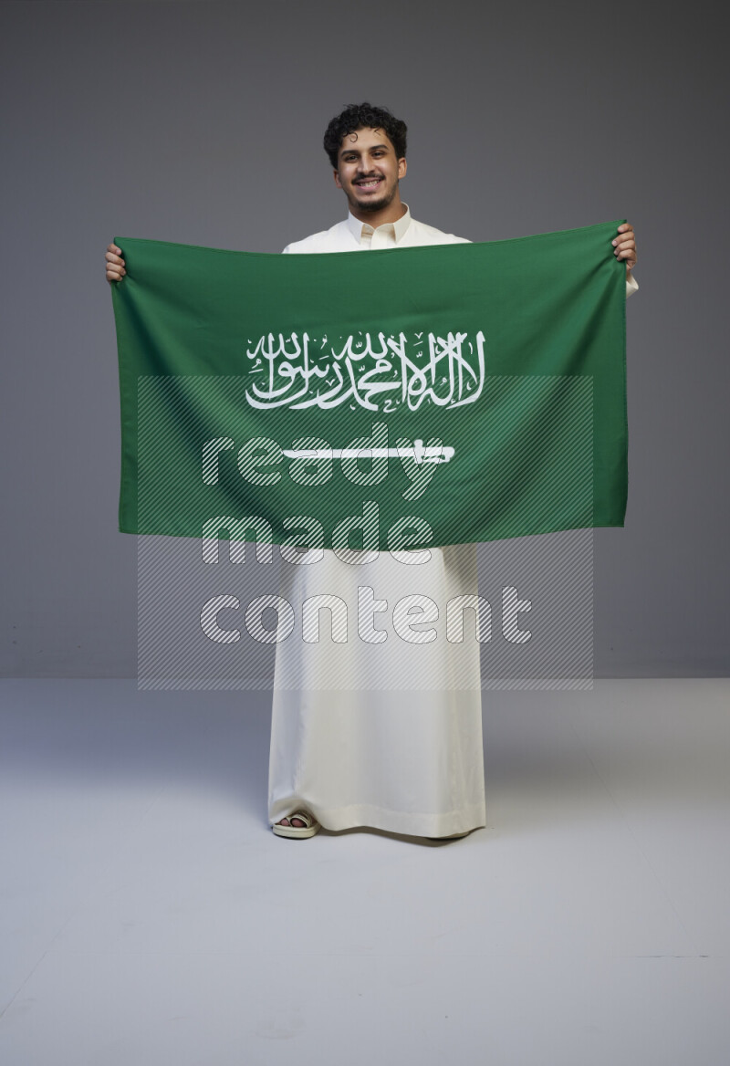 A Saudi man standing wearing thob holding big Saudi flag on gray background