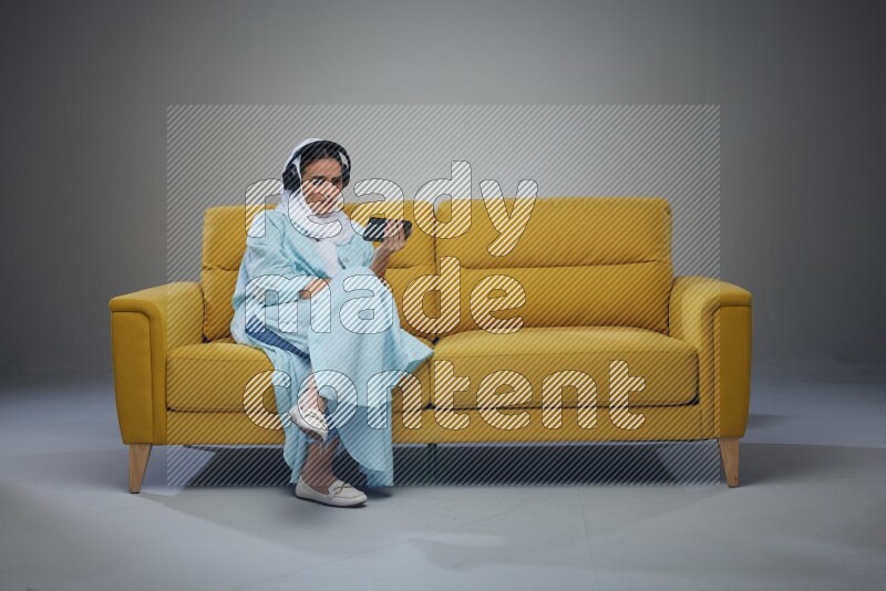 A Saudi woman sitting on a yellow Sofa Watching a Movie wearing Blue Abaya