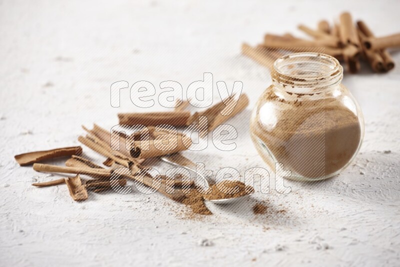 Herbal glass jar full cinnamon powder and a metal spoon surrounded by cinnamon sticks on a white background