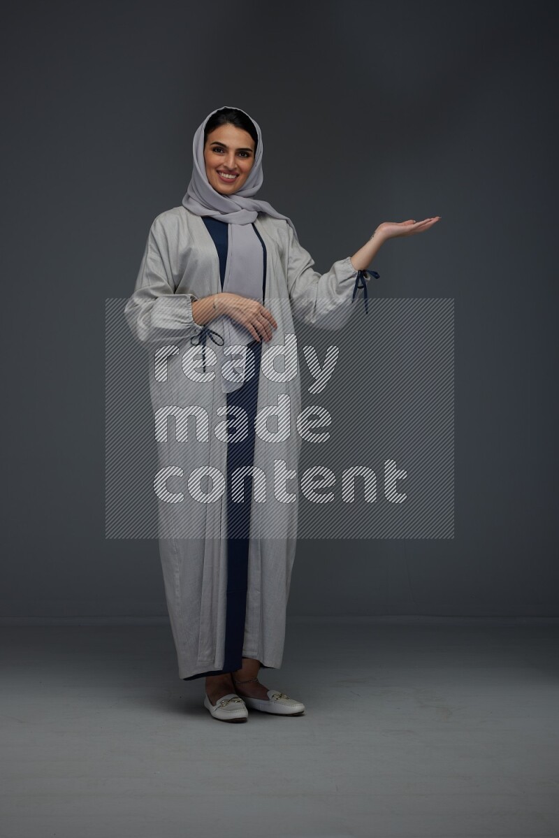 A Saudi woman wearing a light gray Abaya and head scarf standing and pointing in different directions eye level on a grey background