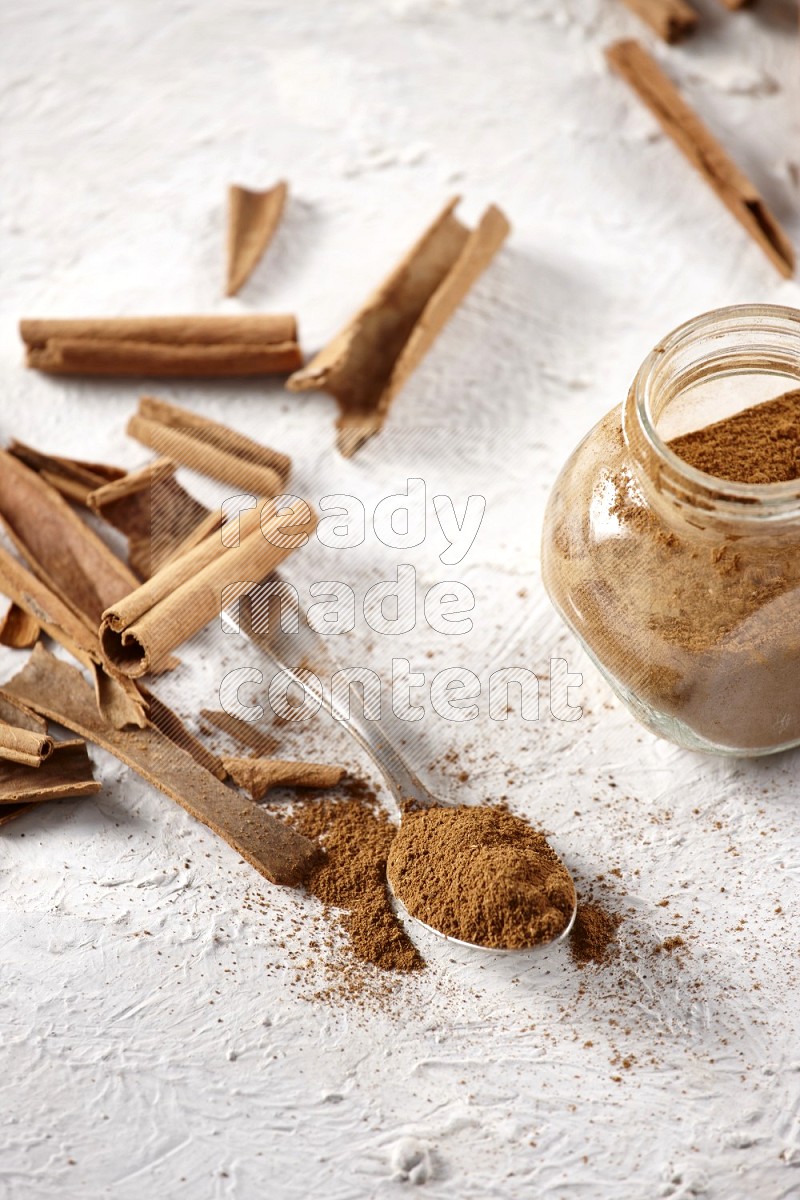 Herbal glass jar full cinnamon powder and a metal spoon surrounded by cinnamon sticks on a white background