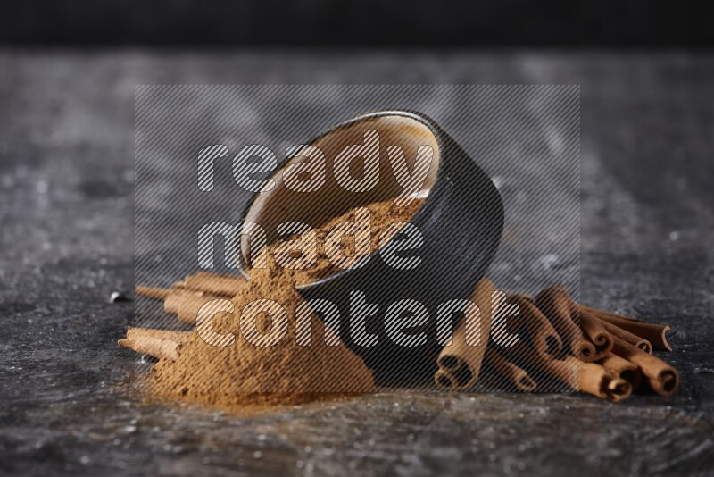 Black pottery bowl over filled with cinnamon powder and cinnamon sticks around the bowl on a textured black background