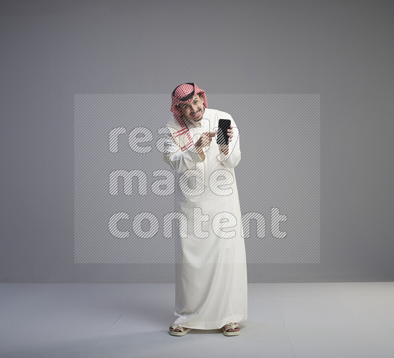 A Saudi man standing wearing thob and red shomag showing phone to camera on gray background