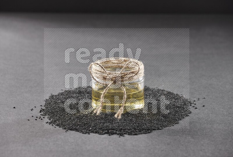 A glass jar full of black seeds oil surrounded by the seeds on a black flooring