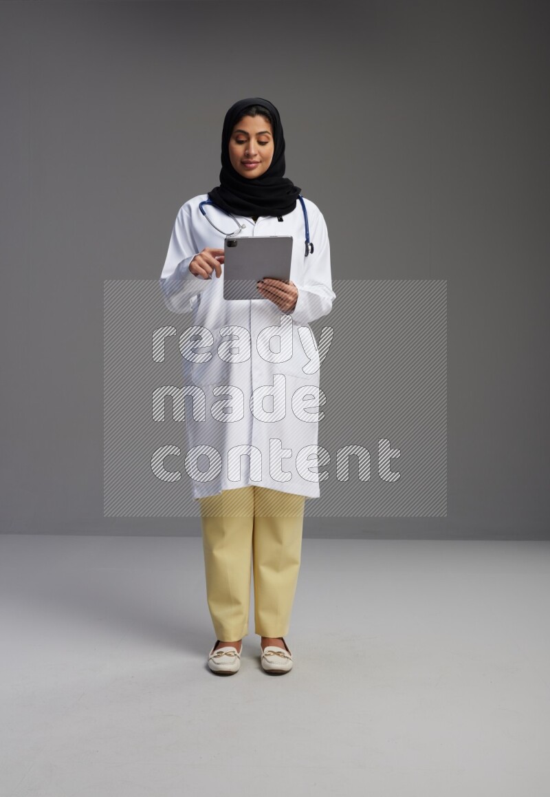 Saudi woman wearing lab coat with stethoscope standing working on tablet on Gray background