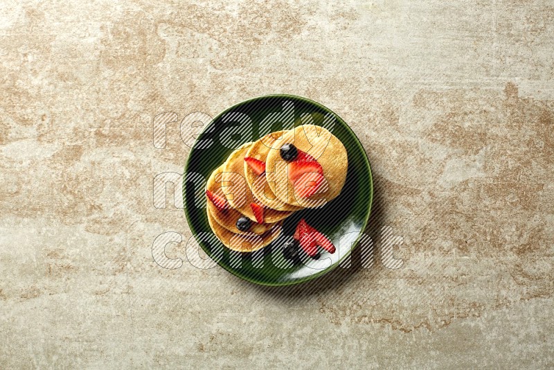 Five stacked mixed berries mini pancakes in a green plate on beige background