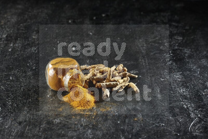 وعاء زجاجي للتوابل ممتلئ ببودرة الكركم مقلوب والبودرة تناثرت منه مع أصابع الكركم المجففة على خلفية سوداء