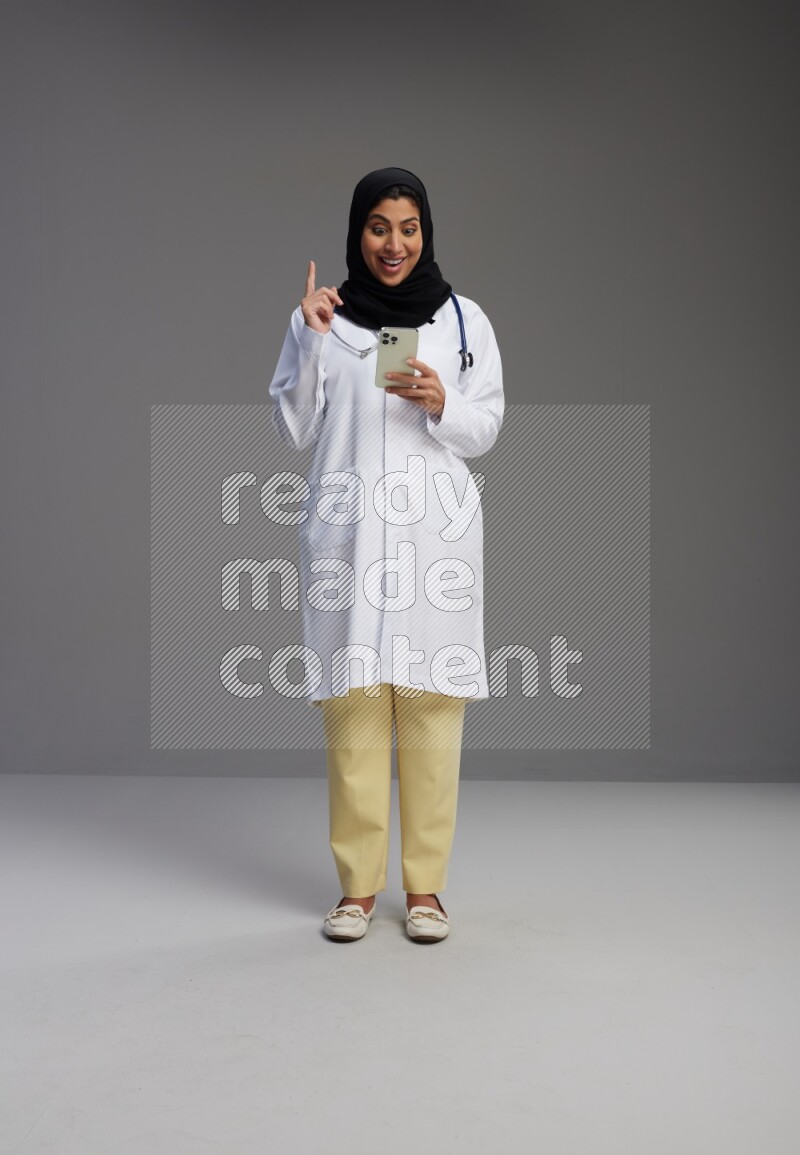 Saudi woman wearing lab coat with stethoscope standing texting on phone on Gray background