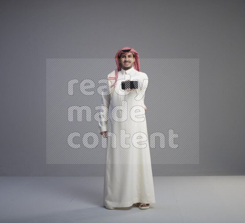 A Saudi man standing wearing thob and red shomag showing phone to camera on gray background